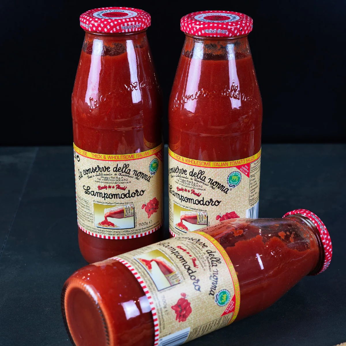 Three glass bottles of Italian tomato sauce with red caps and tan labels on a dark surface.