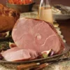 Cooked ham on decorative plate with slices, fruits, garnishes; background has dish and drink.