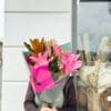 Person holding pink lily bouquet near store window with baskets; face obscured by flowers.