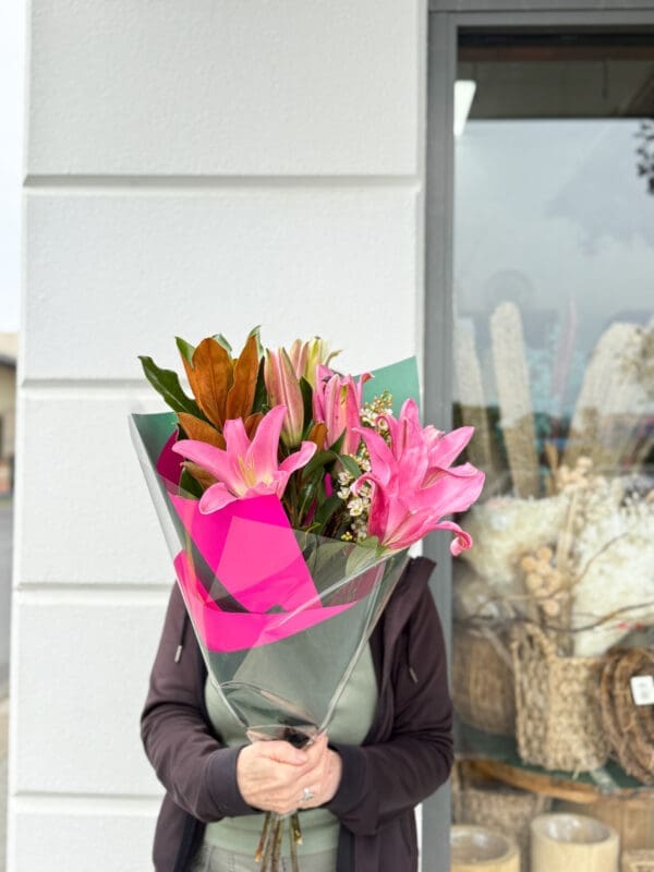 Person holding pink lily bouquet near store window with baskets; face obscured by flowers.