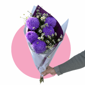 Hand holding bouquet of purple flowers and daisies wrapped in paper with pink background.