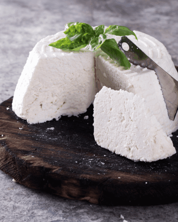 Soft white cheese on board with basil leaves and knife on gray surface.