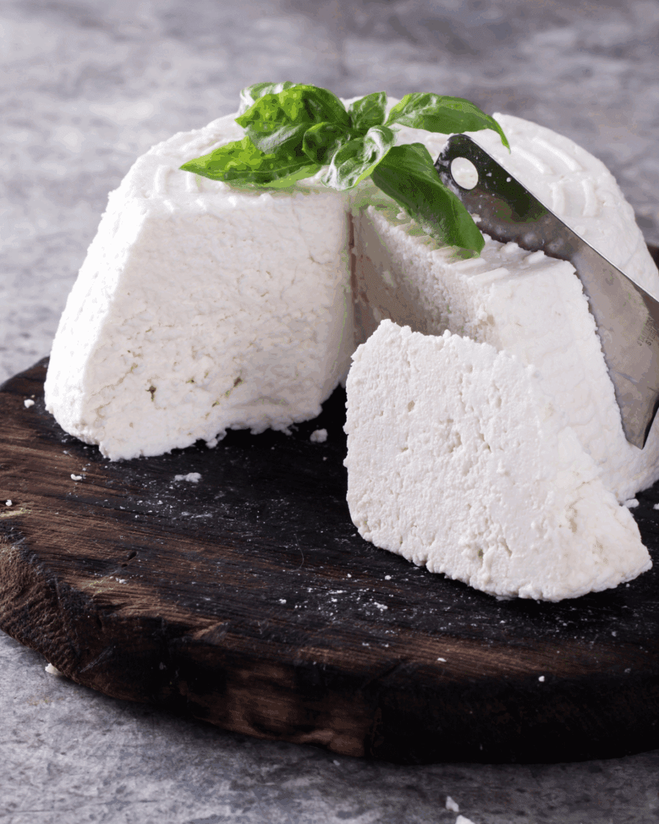 Soft white cheese on board with basil leaves and knife on gray surface.