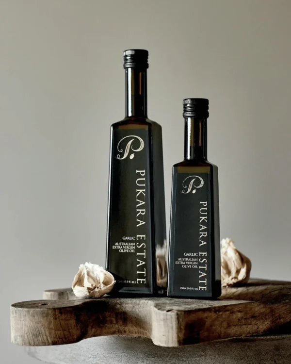 Two Pukara Estate garlic-flavored olive oil bottles on wood, surrounded by garlic cloves.