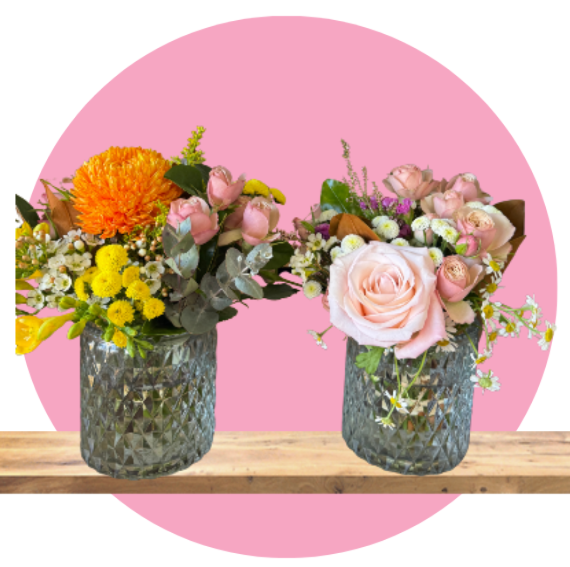 Two floral arrangements in glass vases on wood, with a pink circular backdrop.