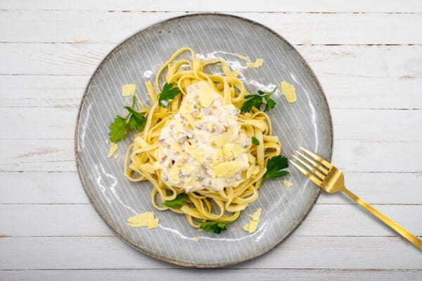 Plate of fettuccine with creamy sauce, parsley, cheese flakes, gold fork on white wood.