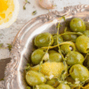 Green olives in ornate dish with lemon peel, thyme, and garlic; halved lemon and clove nearby.