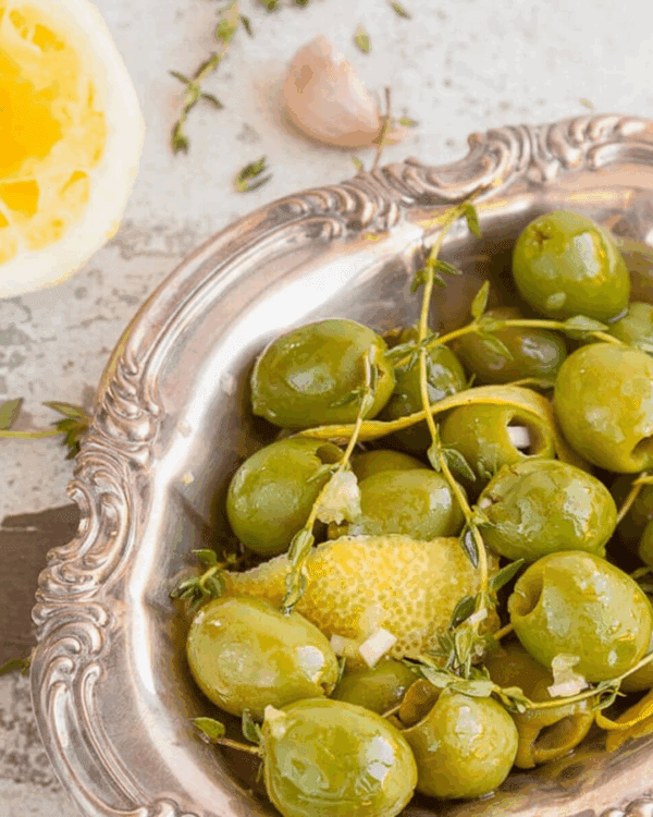 Green olives in ornate dish with lemon peel, thyme, and garlic; halved lemon and clove nearby.