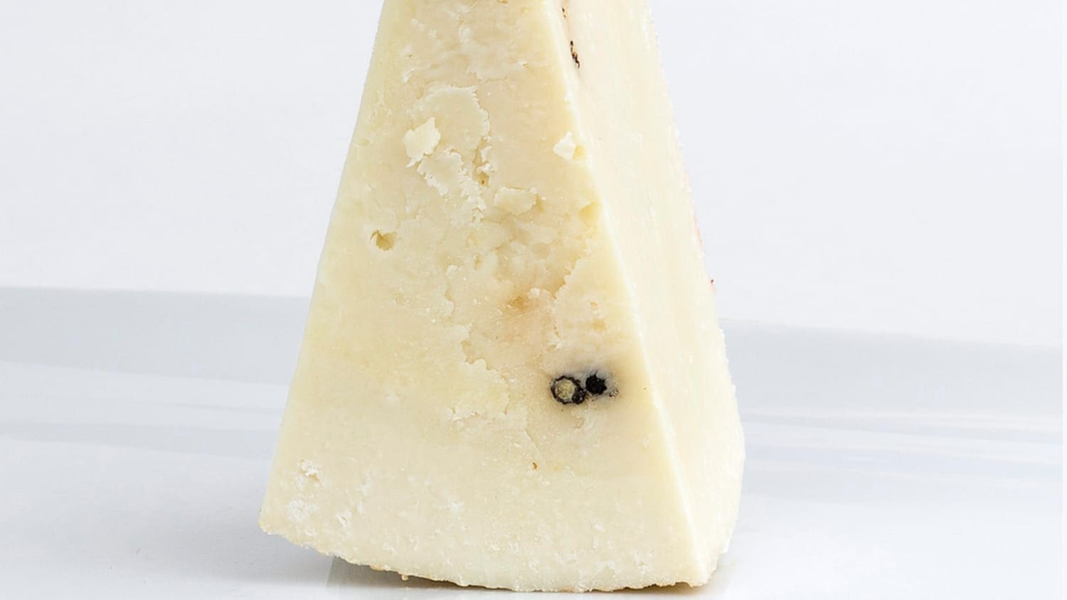 Wedge of hard, aged cheese with crumbly texture and dark spots on off-white surface.