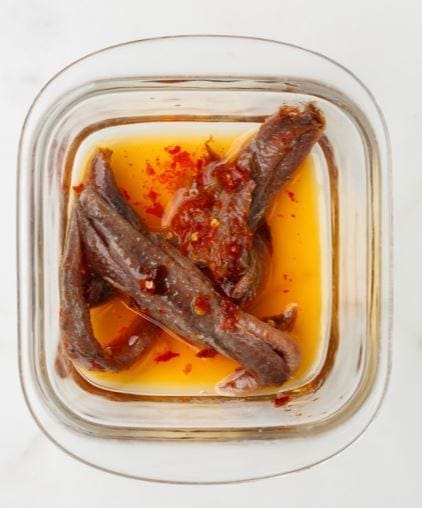 Glass container with anchovy fillets in oil, topped with red chili flakes.