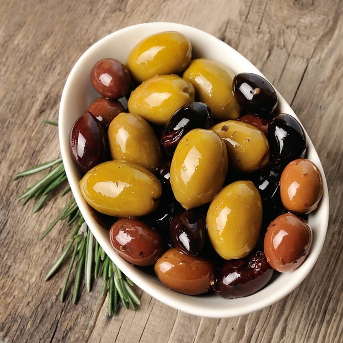 Bowl of assorted olives in green, brown, black with rosemary on wooden surface.