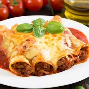 Plate of baked cannelloni with meat, cheese, basil, tomato sauce; tomatoes, olive oil behind.