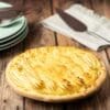 Baked pie with golden crust on wood; green plates, cutlery, napkin, and serving utensil nearby.