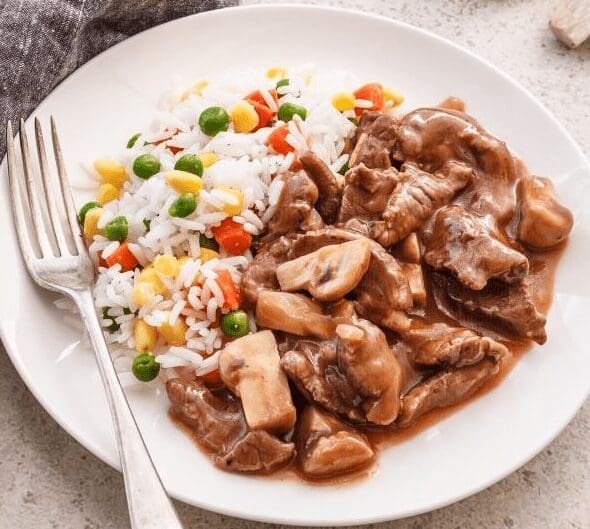 Beef stew with rice, peas, corn, carrots, fork on left, served on a white plate.