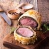Beef Wellington sliced on board with knife, salt, pepper, and rosemary garnish.