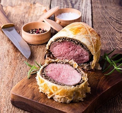 Beef Wellington sliced on board with knife, salt, pepper, and rosemary garnish.