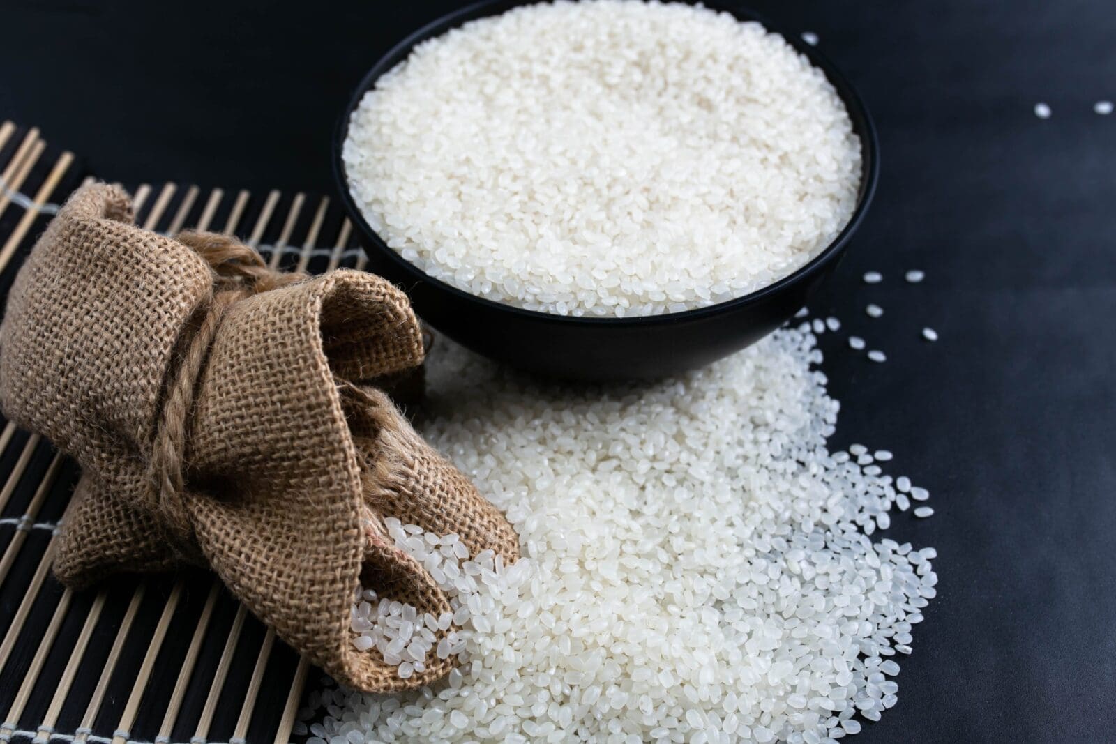 Black bowl of white rice on dark surface, jute sack spilling rice onto bamboo mat.