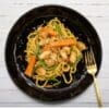 Black plate with noodles, chicken, carrots, green beans, and gold fork on white wooden background.