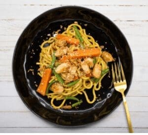 Black plate with noodles, chicken, carrots, green beans, and gold fork on white wooden background.