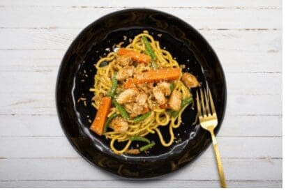 Black plate with noodles, chicken, carrots, green beans, and gold fork on white wooden background.
