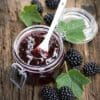 Glass jar of blackberry jam with spoon, fresh blackberries, leaves on wooden surface.