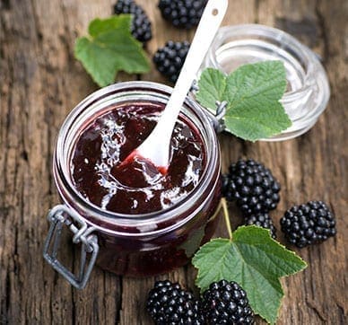 Glass jar of blackberry jam with spoon, fresh blackberries, leaves on wooden surface.
