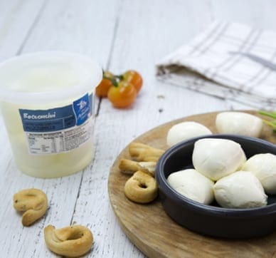 Bowl of bocconcini cheese with bread snacks, tub, tomatoes, and a towel on a wooden board.
