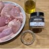 Bowl of chicken, jerk seasoning, spices, and olive oil on wooden surface.