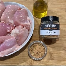 Bowl of chicken, jerk seasoning, spices, and olive oil on wooden surface.