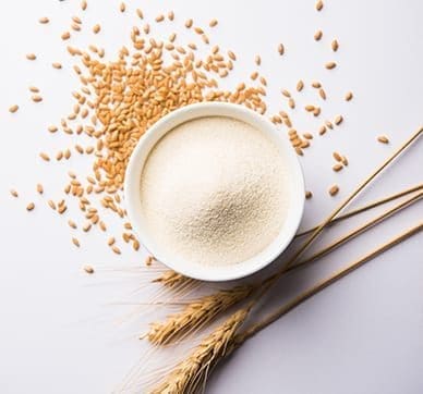 Bowl of flour surrounded by scattered wheat grains and stalks on a white surface.