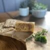 Butter roll on wooden board with herbs and two seeded crackers, blurred potted plants behind.