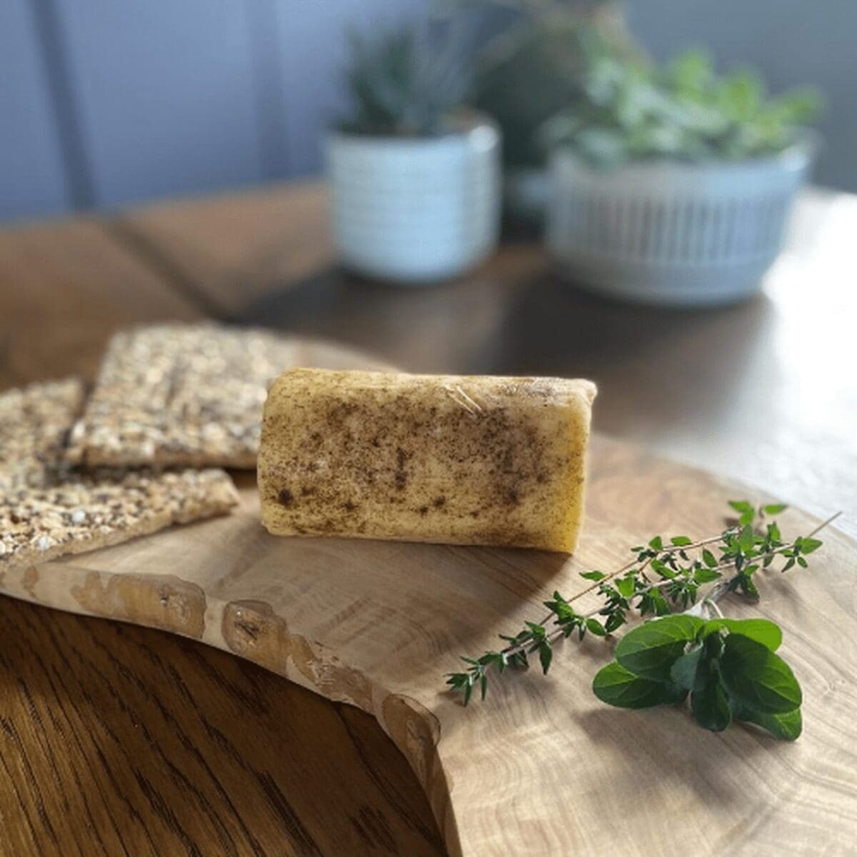 Butter roll on wooden board with herbs and two seeded crackers, blurred potted plants behind.