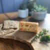 Cheese with herbs, seeded crackers, and green herbs on board; potted plants in background.