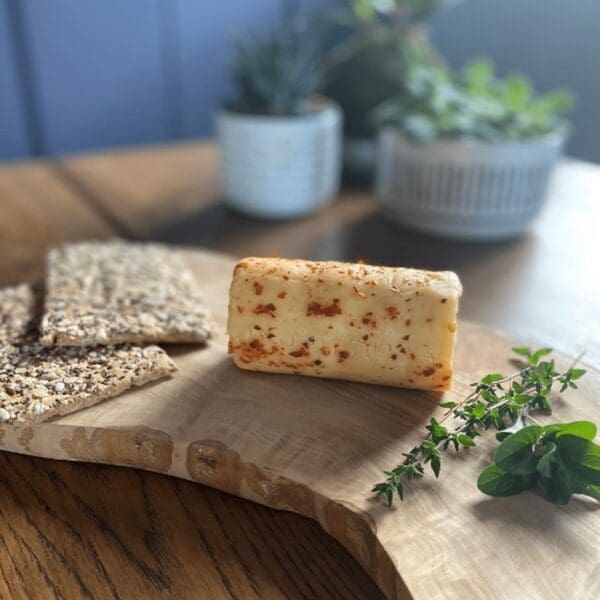 Cheese with herbs, seeded crackers, and green herbs on board; potted plants in background.