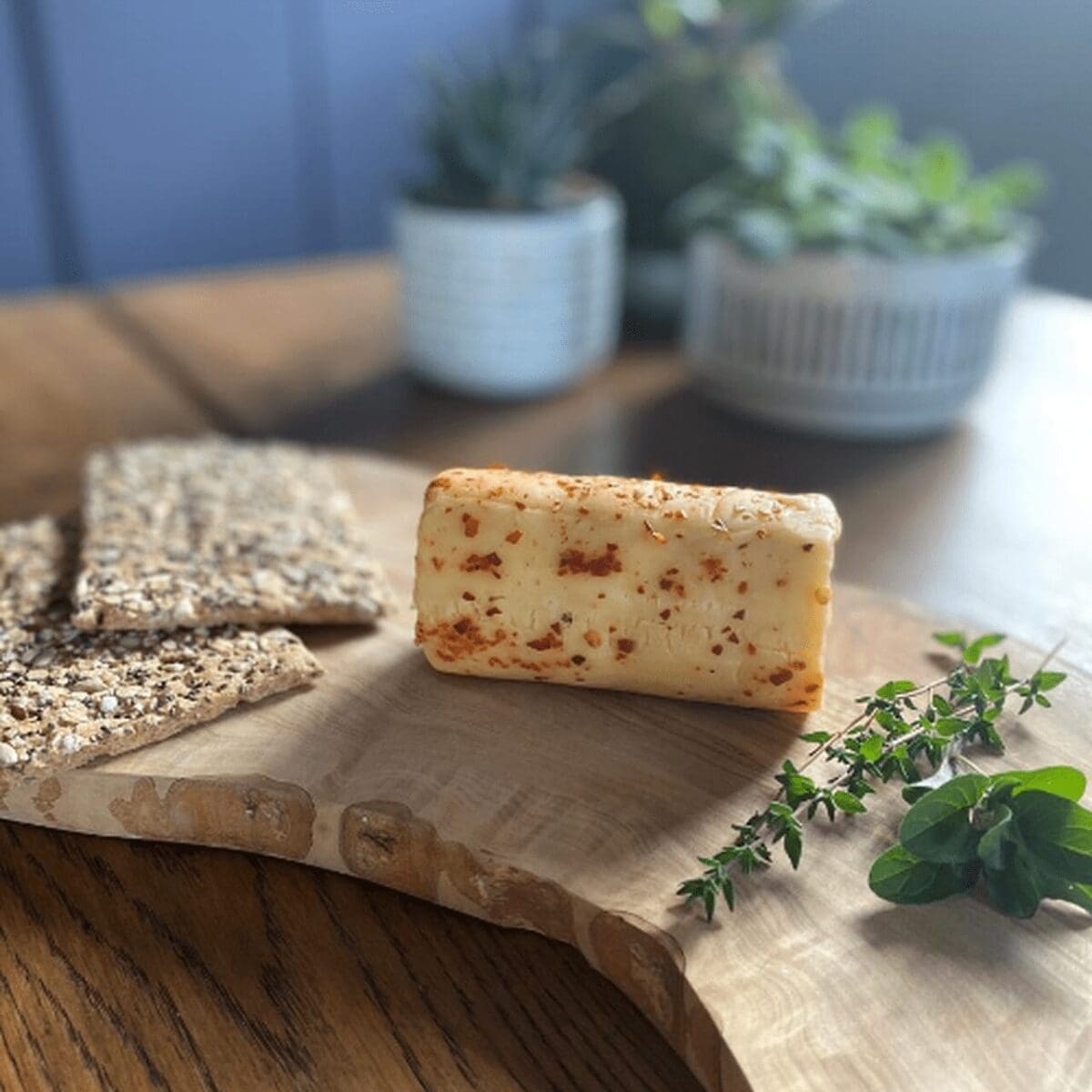 Cheese with herbs, seeded crackers, and green herbs on board; potted plants in background.