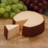 Round cheese wheel with a wedge cut, placed on wood board with green and red grapes behind.