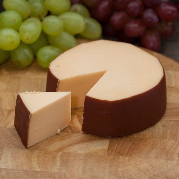 Round cheese wheel with a wedge cut, placed on wood board with green and red grapes behind.
