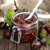 Jar of cherry jam with spoon, metal clasp on wood, surrounded by fresh cherries and leaves.