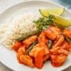 White plate with rice, chicken in red sauce, green vegetables, cilantro, and lime wedges.