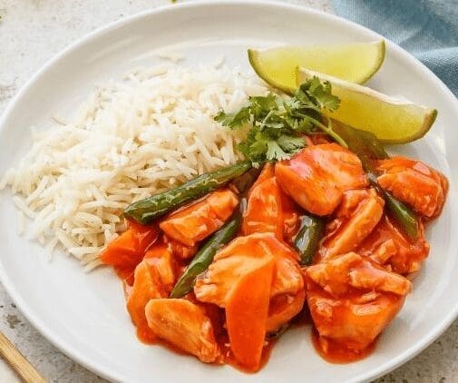 White plate with rice, chicken in red sauce, green vegetables, cilantro, and lime wedges.