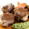 Cooked meat pieces in sauce with green puree, carrots, and green beans in the background.