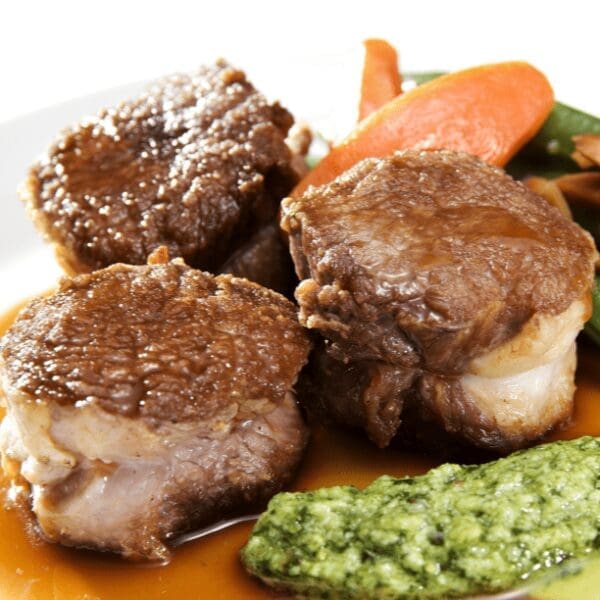 Cooked meat pieces in sauce with green puree, carrots, and green beans in the background.