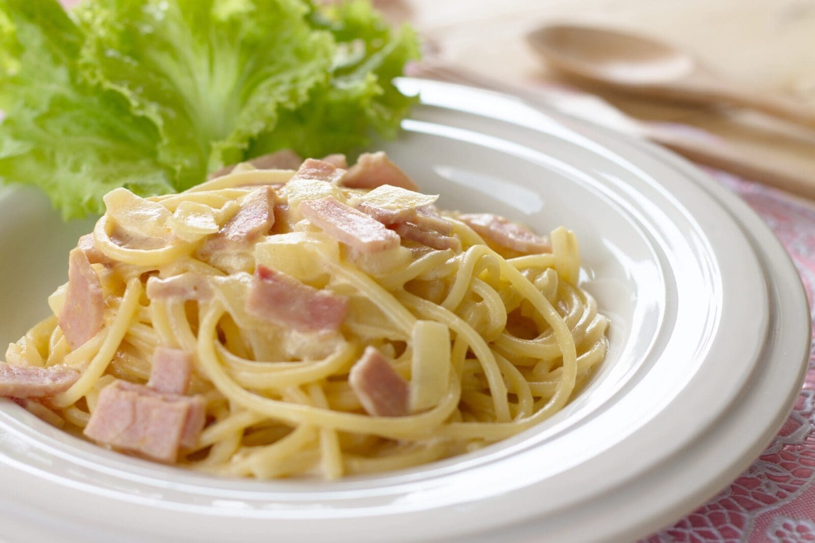 Pasta with creamy sauce and bacon, garnished with lettuce on a white plate, soft focus utensils.