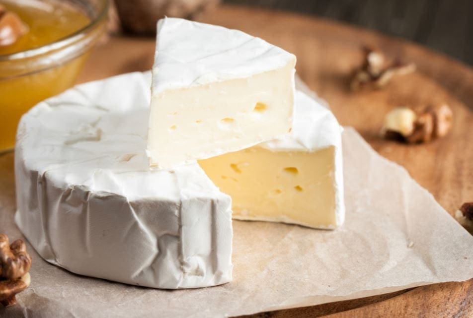 Round creamy cheese on parchment with a wedge cut out, walnuts, and honey on a wooden board.