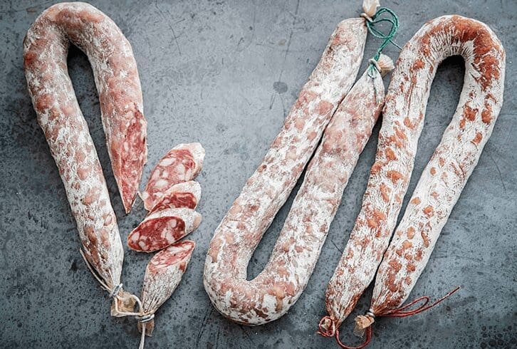 Cured sausages on gray surface, some sliced to show marbled insides and tied with strings.