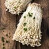 Cluster of enoki mushrooms on wood, garnished with chopped green herbs.