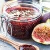 Glass jar of fig jam, half fig, wooden spoon with jam on wooden surface.