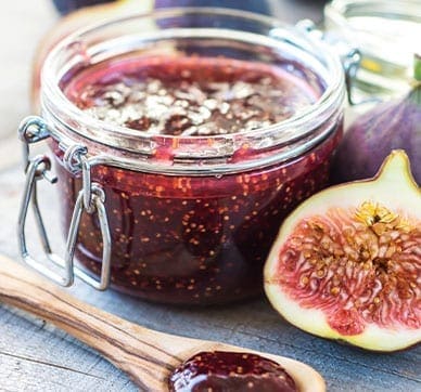 Glass jar of fig jam, half fig, wooden spoon with jam on wooden surface.