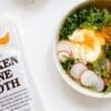 Free Range Chicken Bone Broth package beside a soup with greens, radishes, cream, and meat.