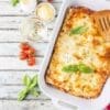 Freshly baked lasagna in dish with basil, cherry tomatoes, cheese, wine, and checkered cloth.
